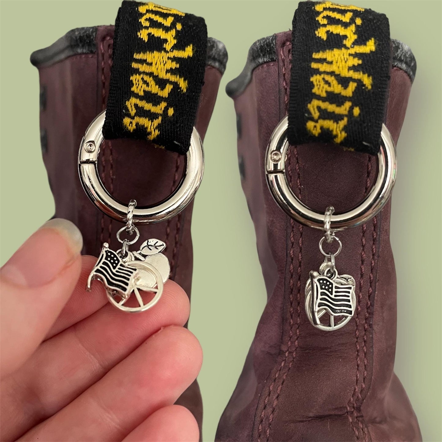 Close-up of handmade silver boot charms with American flag, circle with peace sign, and apple designs clipped onto black boot tabs