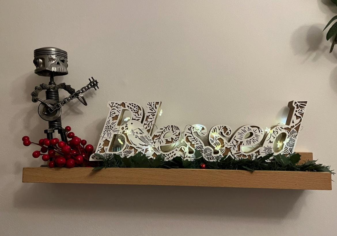 Scrap metal banjo player sculpture made from nuts, bolts, and automotive parts — steampunk-inspired handmade art piece for banjo enthusiasts and collectors, displayed on wooden shelf with red berries and lit word sign saying blessed.