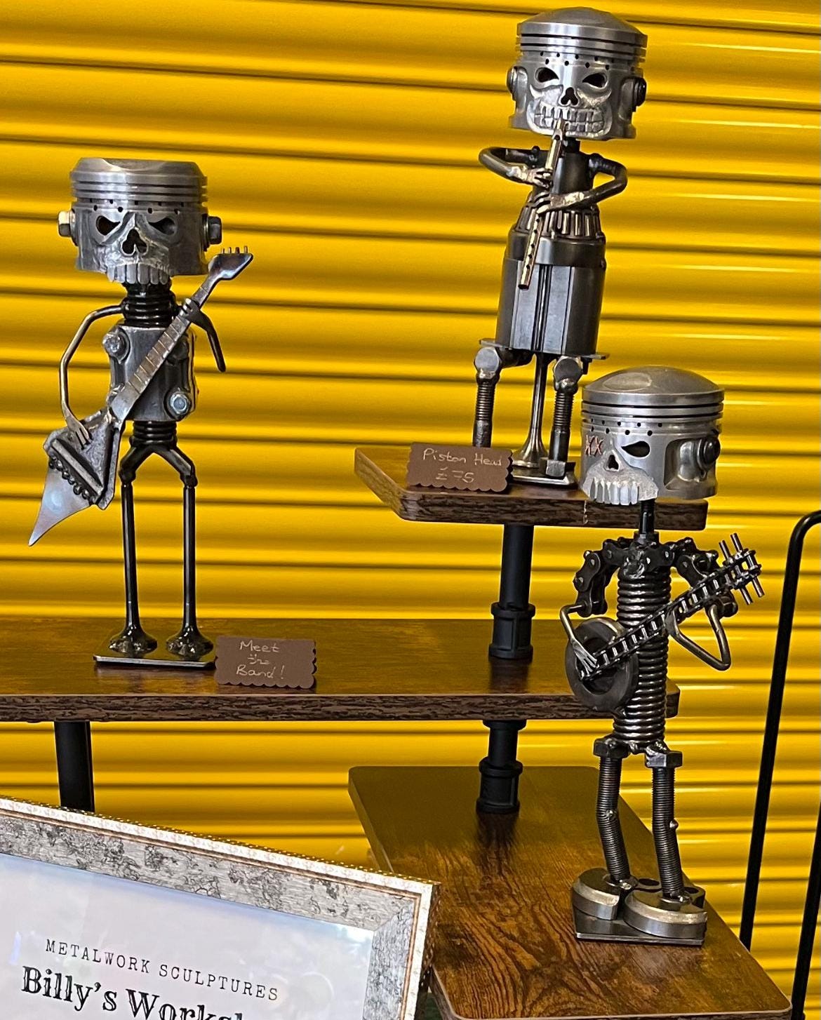 Trio of handmade metal musician sculptures crafted from recycled automotive parts including pistons, bolts, and spanners, displayed against a yellow background — quirky scrap metal art band perfect for music lovers, steampunk collectors, or industrial decor.