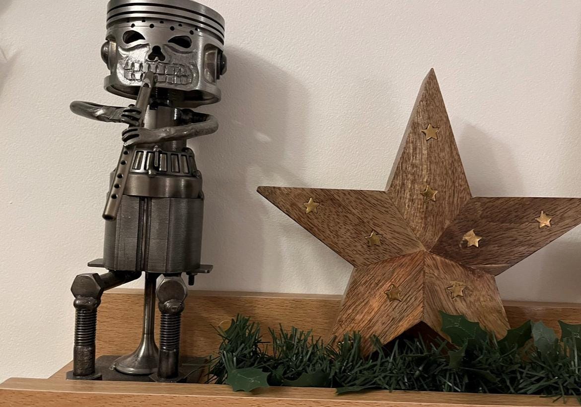 Upcycled metal tin whistle player sculpture with piston skull head, crafted from recycled motorbike parts — industrial-style music-themed home decor or musician gift, displayed on wooden shelf with wooden star.