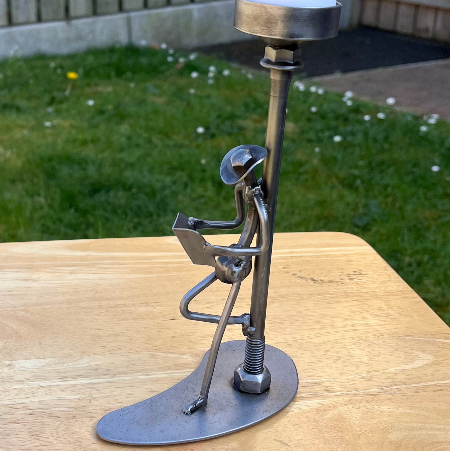 Scrap metal sculpture of a man reading a newspaper while leaning against a lamp post, which doubles as a tealight holder. Handcrafted from upcycled nuts, nails, and other metal parts. Functional industrial art for home or office decor. Facing left.