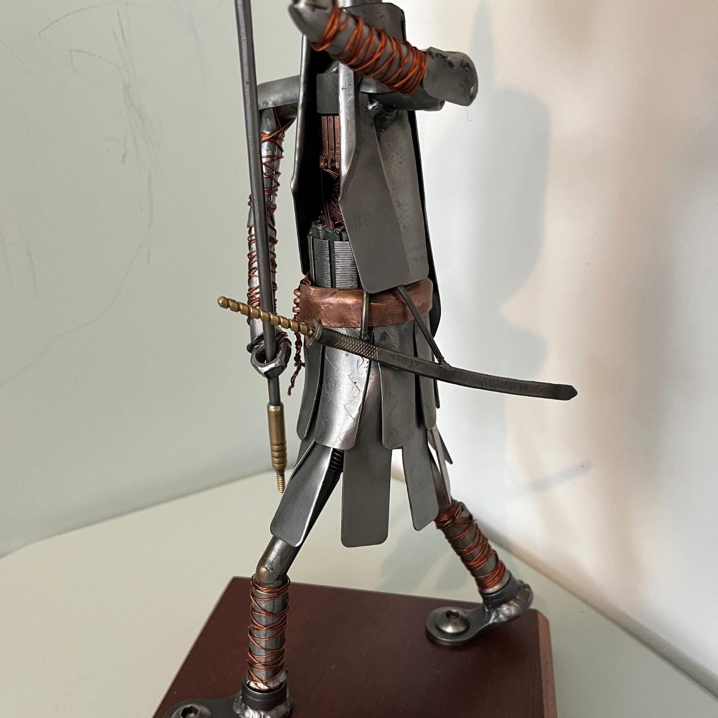 Samurai warrior sculpture made from a piston head, featuring copper and steel armor, a wooden base, and a sword. Close up of lower body.