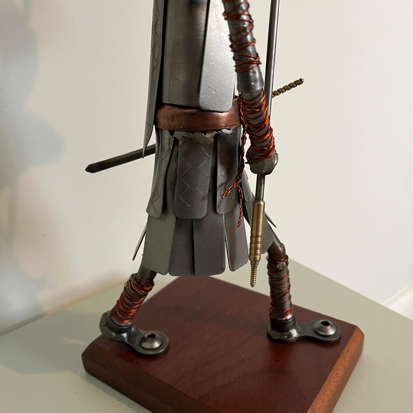 Samurai warrior sculpture made from a piston head, featuring copper and steel armor, a wooden base, and a sword. Close up of lower armour.