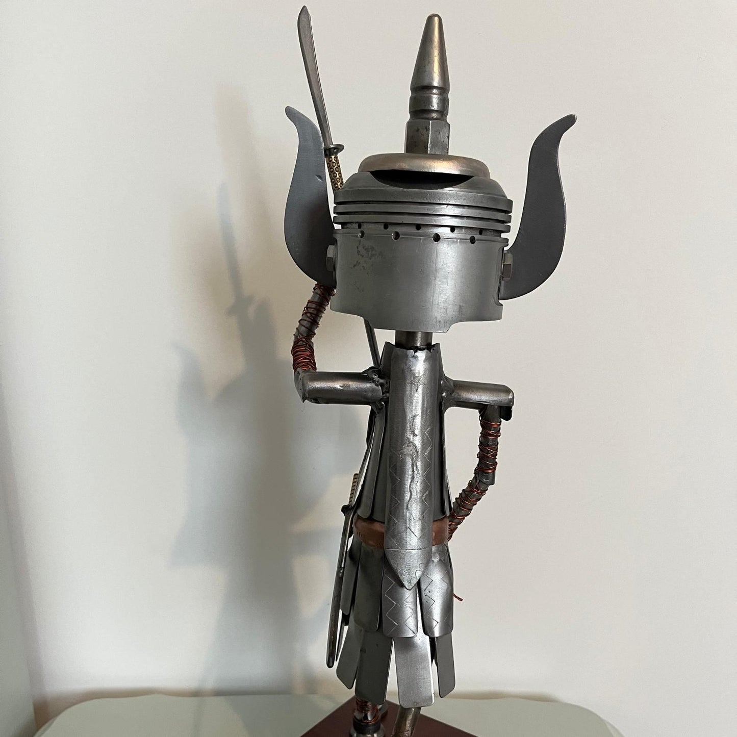 Samurai warrior sculpture made from a piston head, featuring copper and steel armor, a wooden base, and a sword. View of back.
