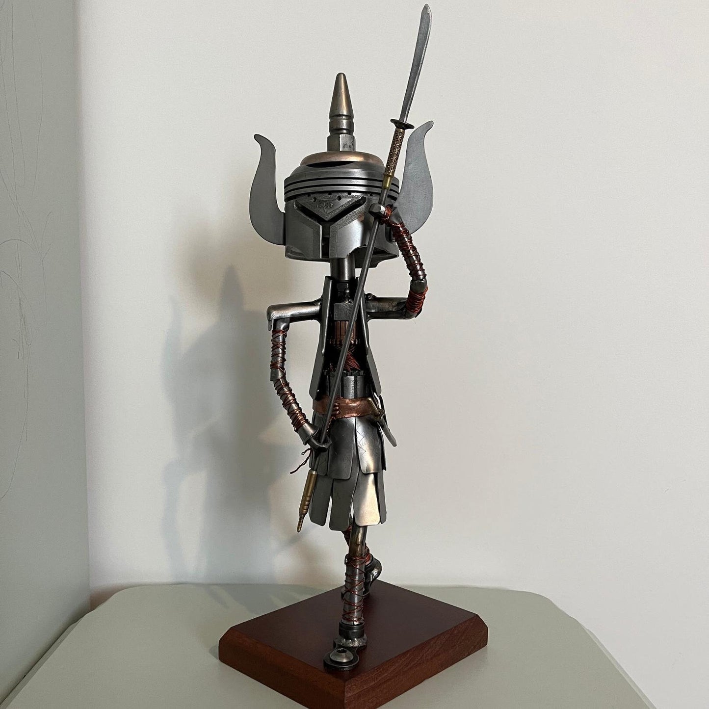 Samurai warrior sculpture made from a piston head, featuring copper and steel armor, a wooden base, and a sword. A unique blend of mechanical art and traditional Japanese warrior aesthetics.