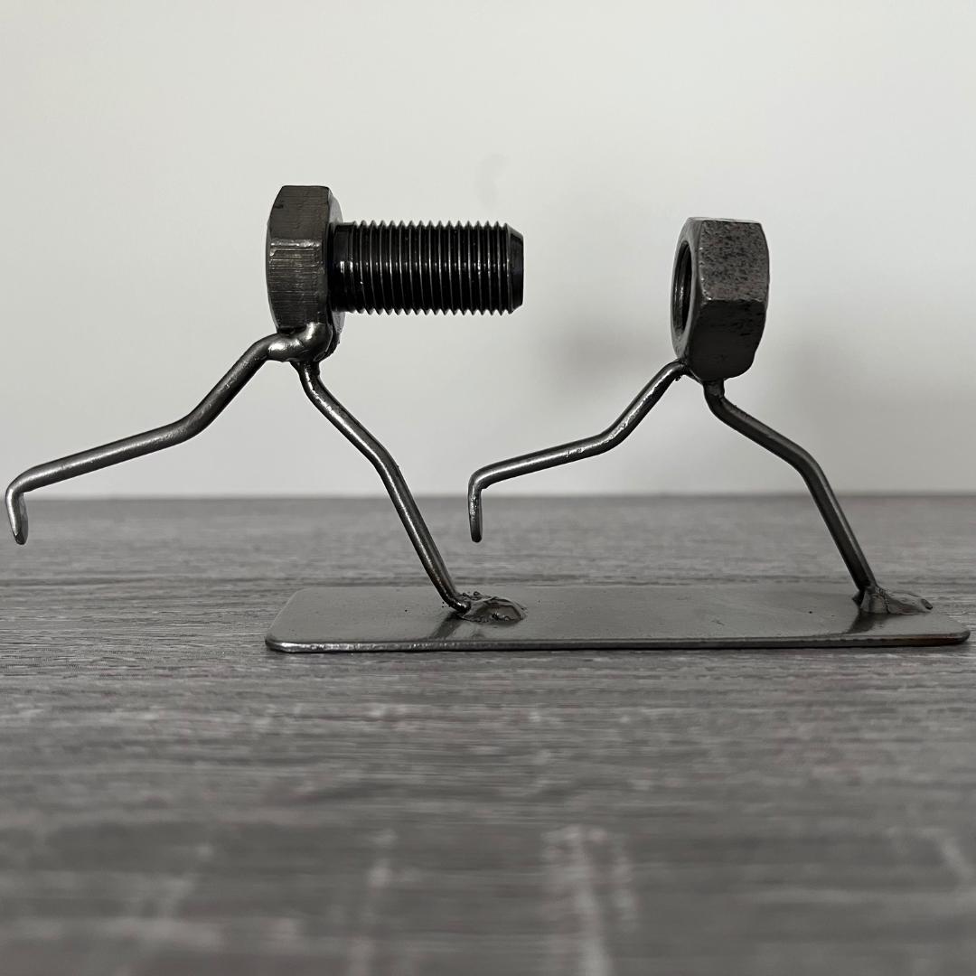 Whimsical sculpture of a bolt chasing a nut, symbolising the playful pursuit of connection. Ideal for tinkerers, engineers, and lovers of metal art and puns. Side view.