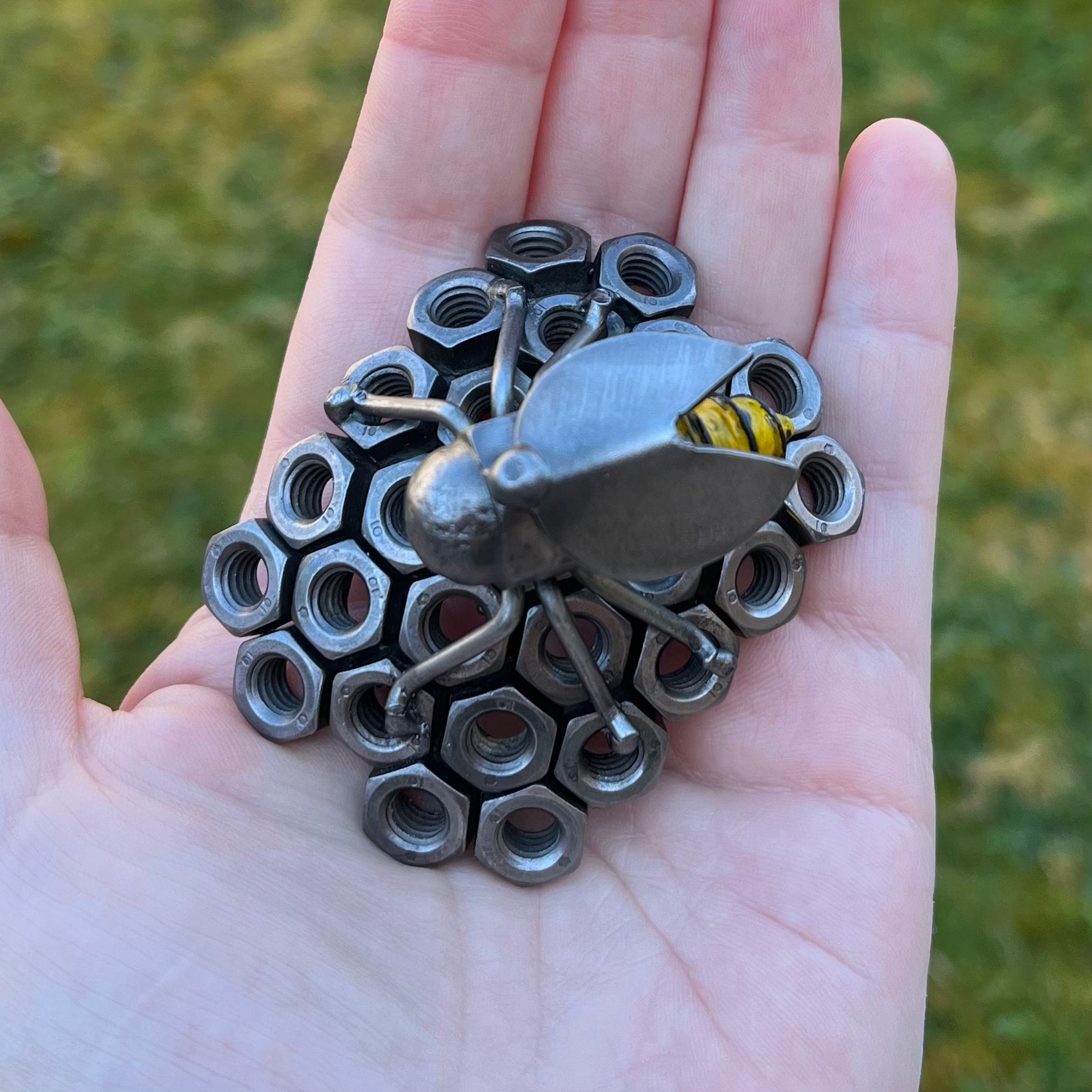 Handmade scrap metal bee sculpture with a screw body and hex nut honeycomb base, unique industrial decor or gift for bee lovers, held by a woman's hand, top view.