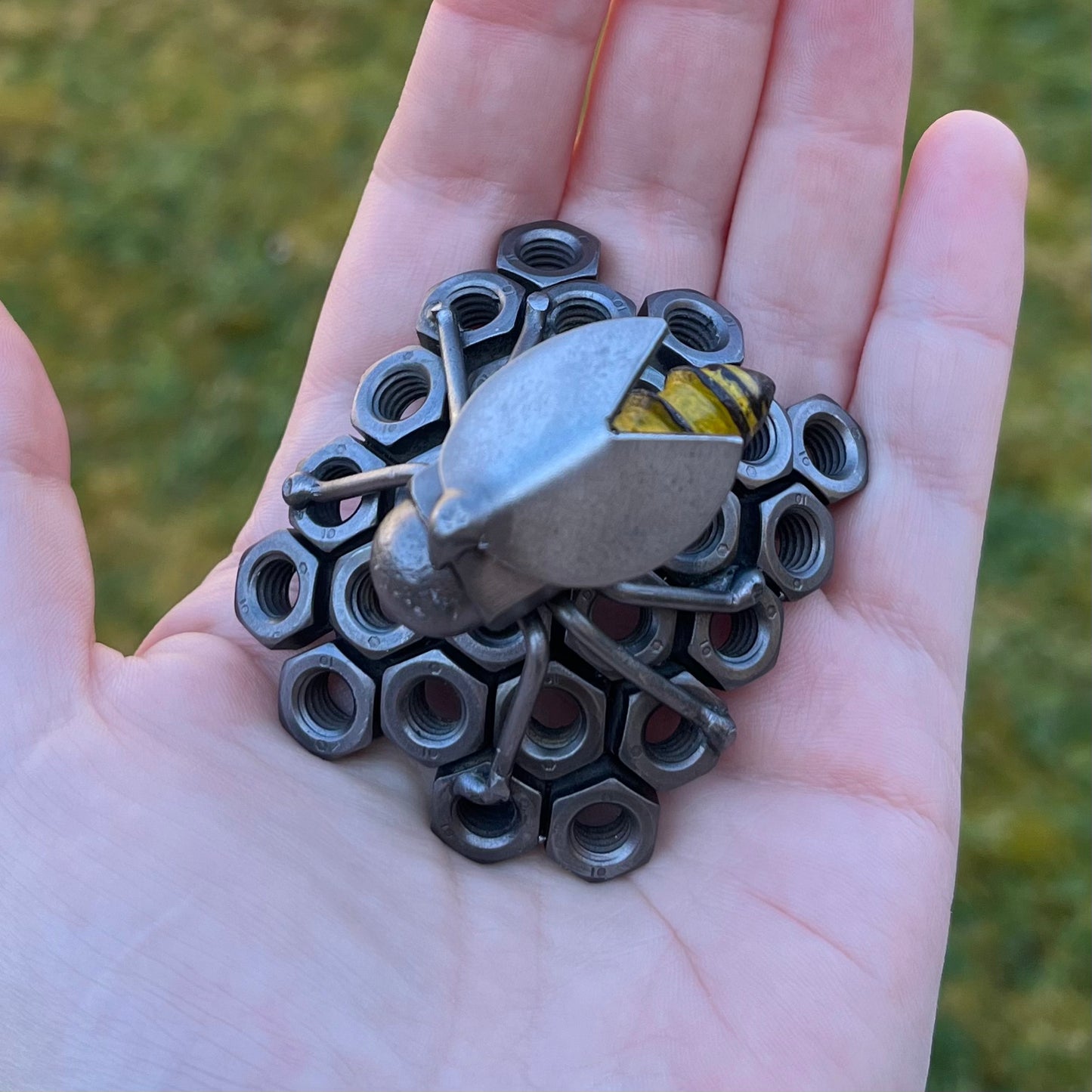 Handmade scrap metal bee sculpture with a screw body and hex nut honeycomb base, unique industrial decor or gift for bee lovers, held by a woman's hand, top view.