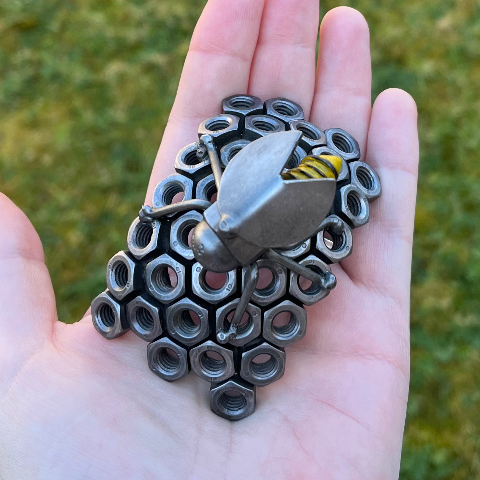 Handmade scrap metal bee sculpture with a screw body and hex nut honeycomb base, unique industrial decor or gift for bee lovers, held by a woman's hand, top view.