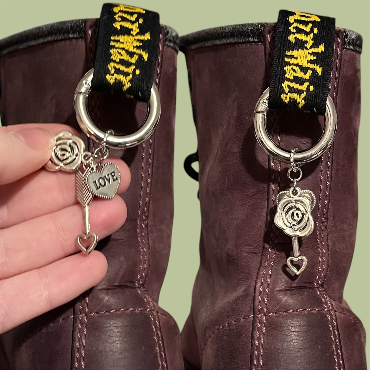 Close-up of handmade silver boot charms with rose, cupid's arrow, and solid heart with love text designs clipped onto black boot tabs